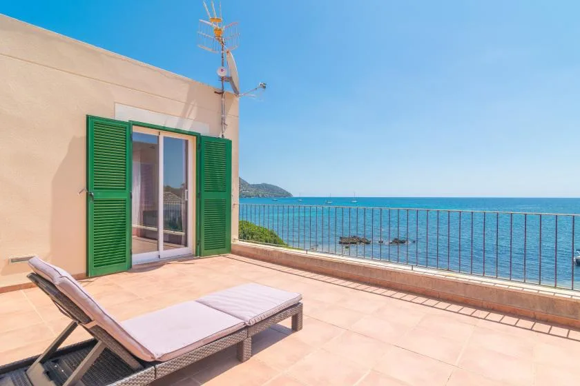A sunlit terrace overlooks the sea, with a lounge chair beside green shutters.