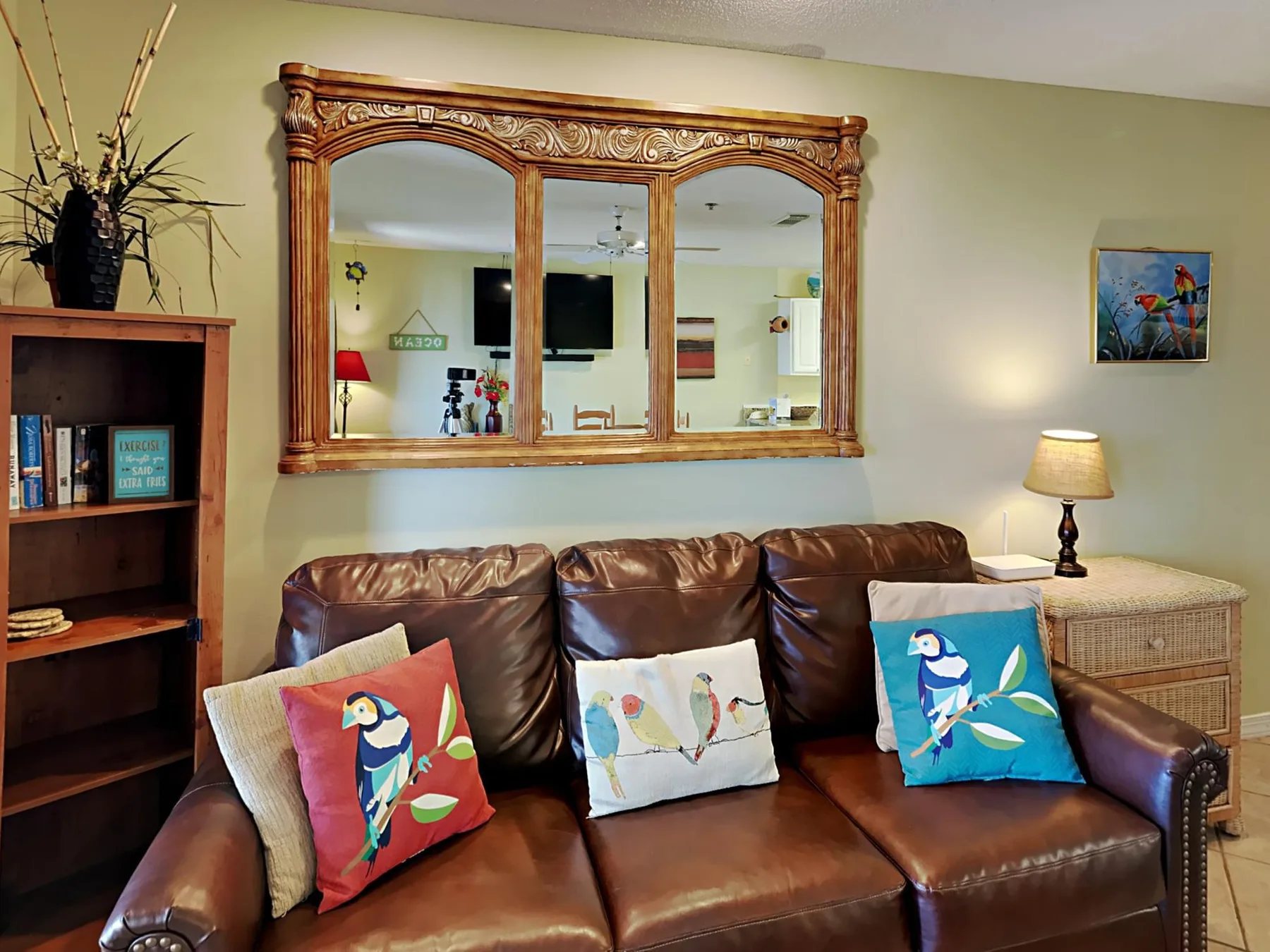 A living room with brown seating, colorful bird cushions, and a large wall mirror.