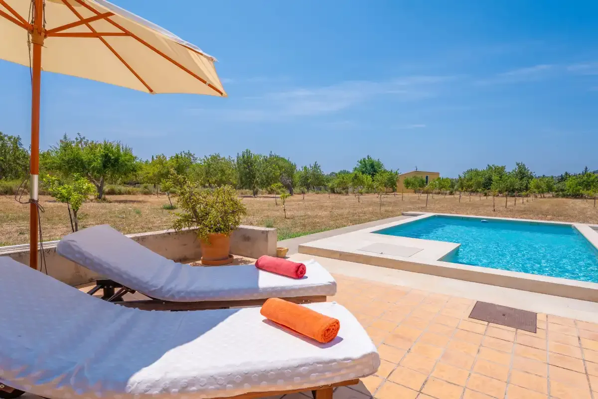 A tiled poolside patio has lounge seating beneath an umbrella beside a swimming pool.