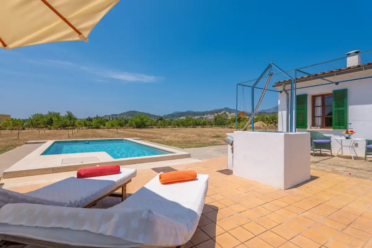 A sunny pool terrace with lounge seating beside a white house.