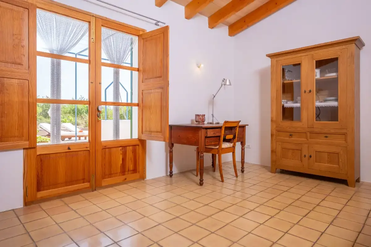 A bright room with wooden window shutters, a writing desk, and a display cabinet.