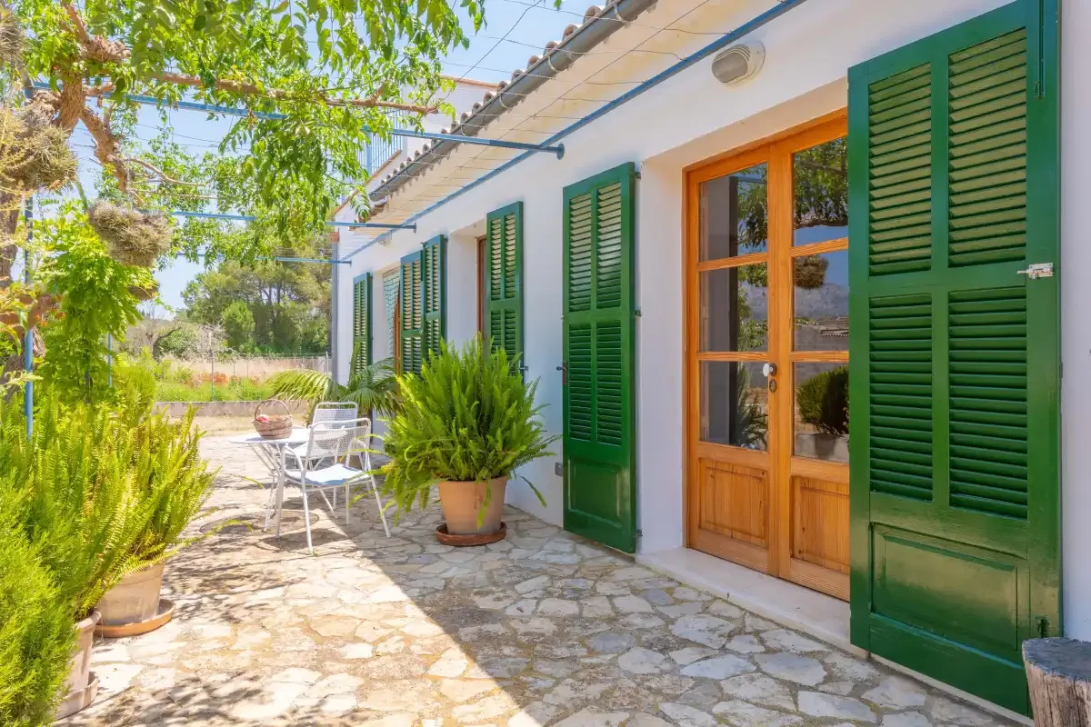 A white house exterior opens onto a stone patio with green shutters and outdoor seating.