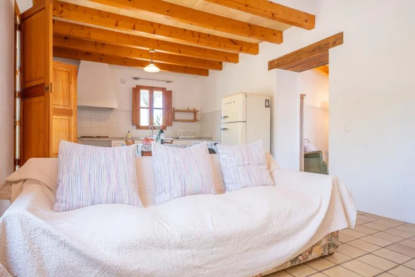 A living area with a large sofa faces an open kitchen beneath exposed wooden beams.