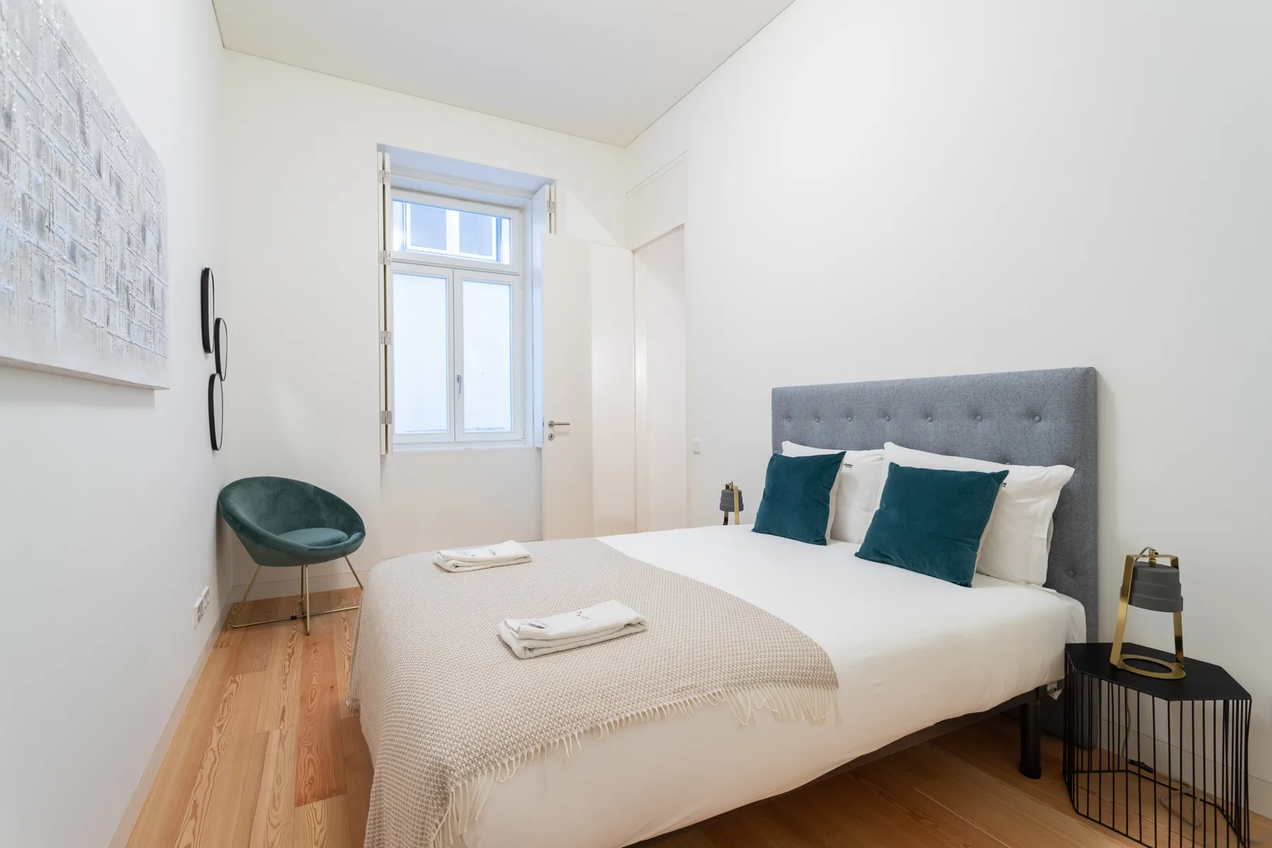 A bright bedroom with a bed, teal cushions, and a window.