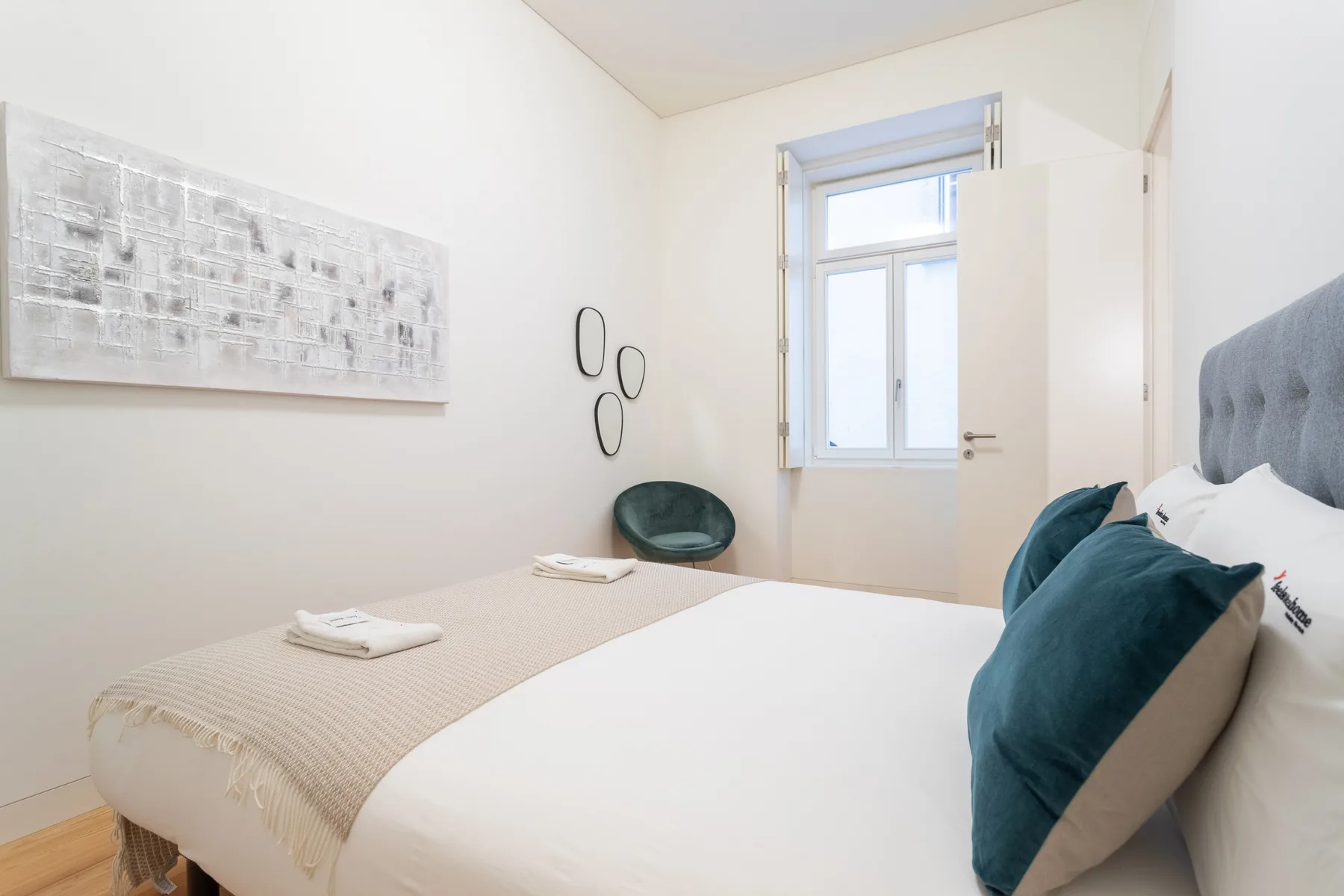 A bright bedroom with a bed, teal cushions, and a window.