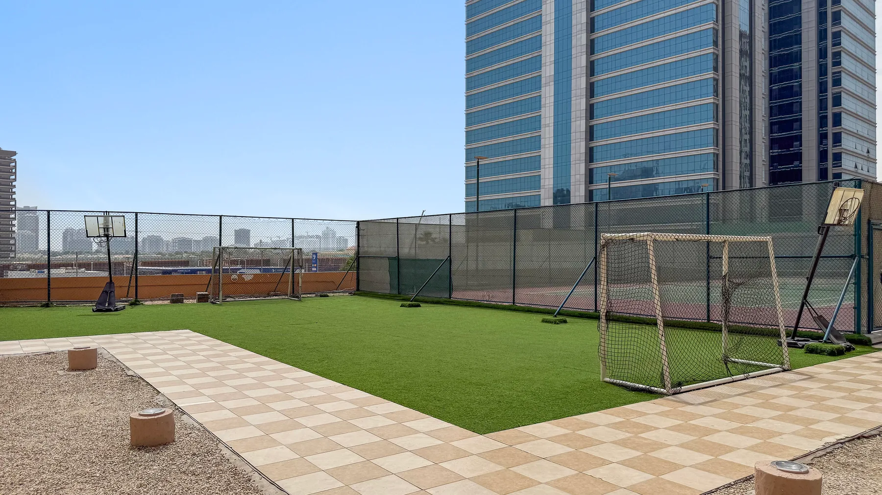 An outdoor sports court with green turf, goals, and tall buildings in the background.