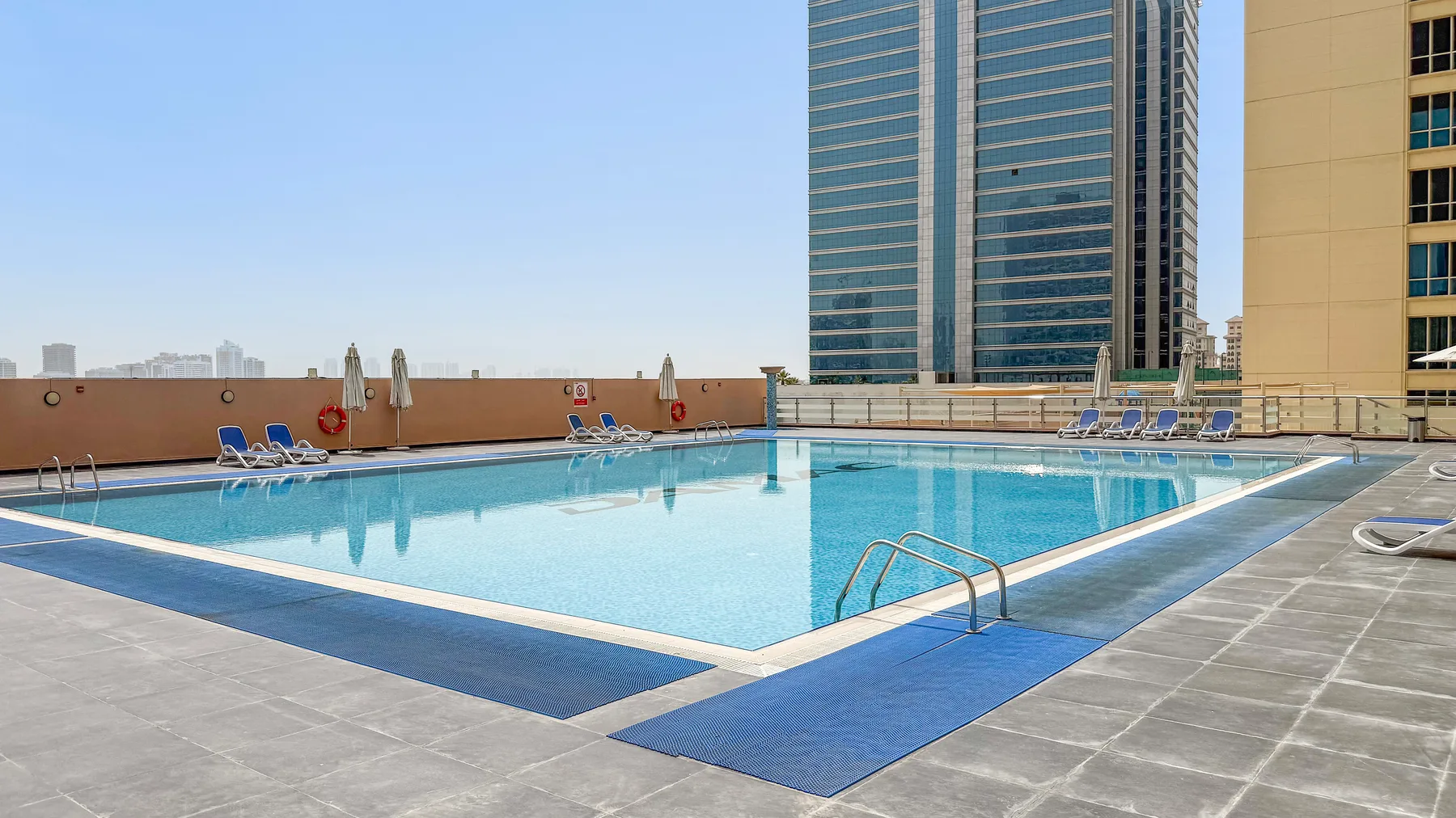 A rooftop swimming pool sits beside a tall modern building under a clear sky.