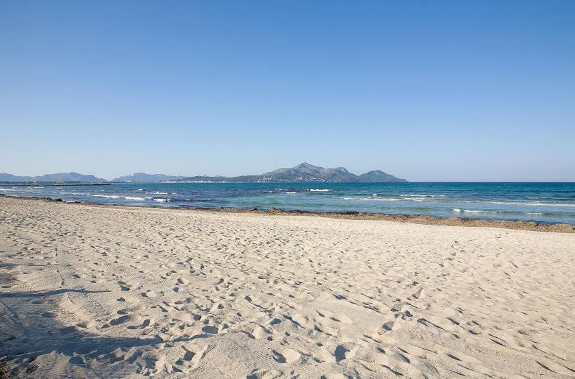 A sandy beach stretches along calm blue water, with distant mountains on the horizon.