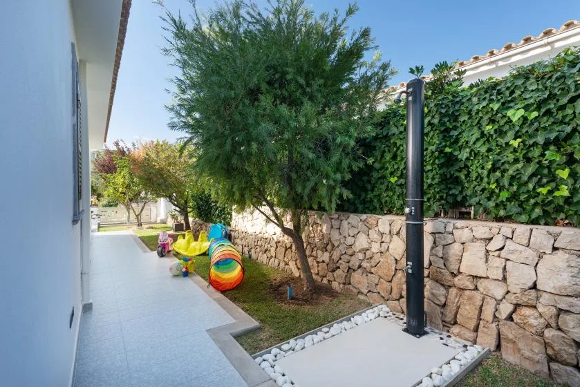 A paved outdoor walkway beside a stone wall, with a leafy tree and colorful play equipment.