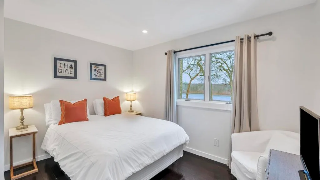 Un dormitorio luminoso con una cama blanca y una ventana con vistas a árboles y agua.
