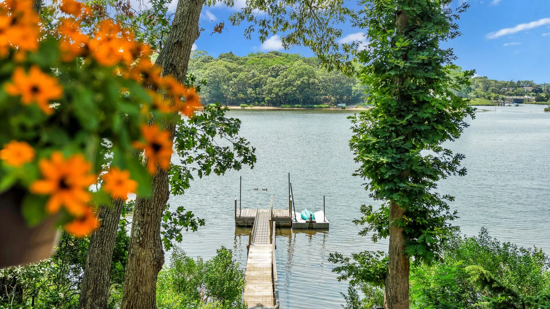 Un muelle de madera se adentra en un lago tranquilo, enmarcado por árboles y flores naranjas.