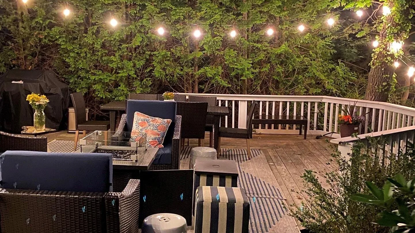 Una terraza exterior con asientos, una mesa de comedor y luces colgantes entre árboles frondosos.