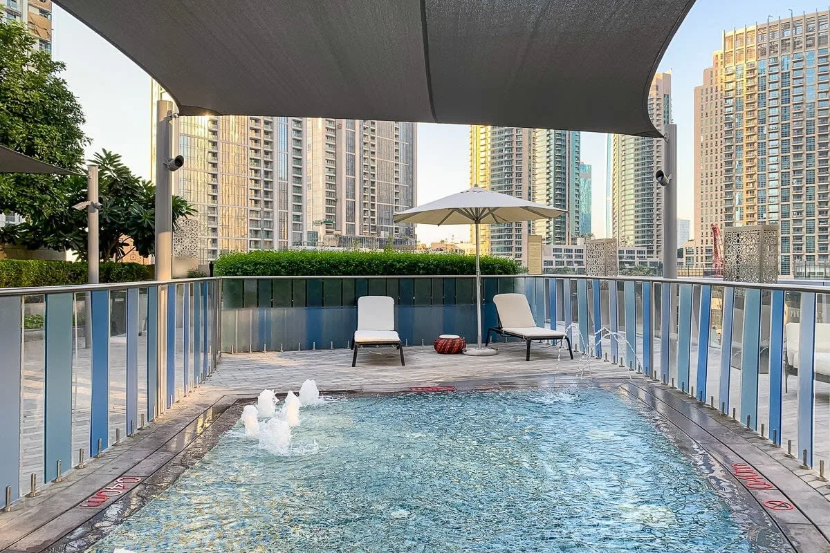 Una piscina al aire libre está bajo un toldo, con asientos y edificios altos al fondo.