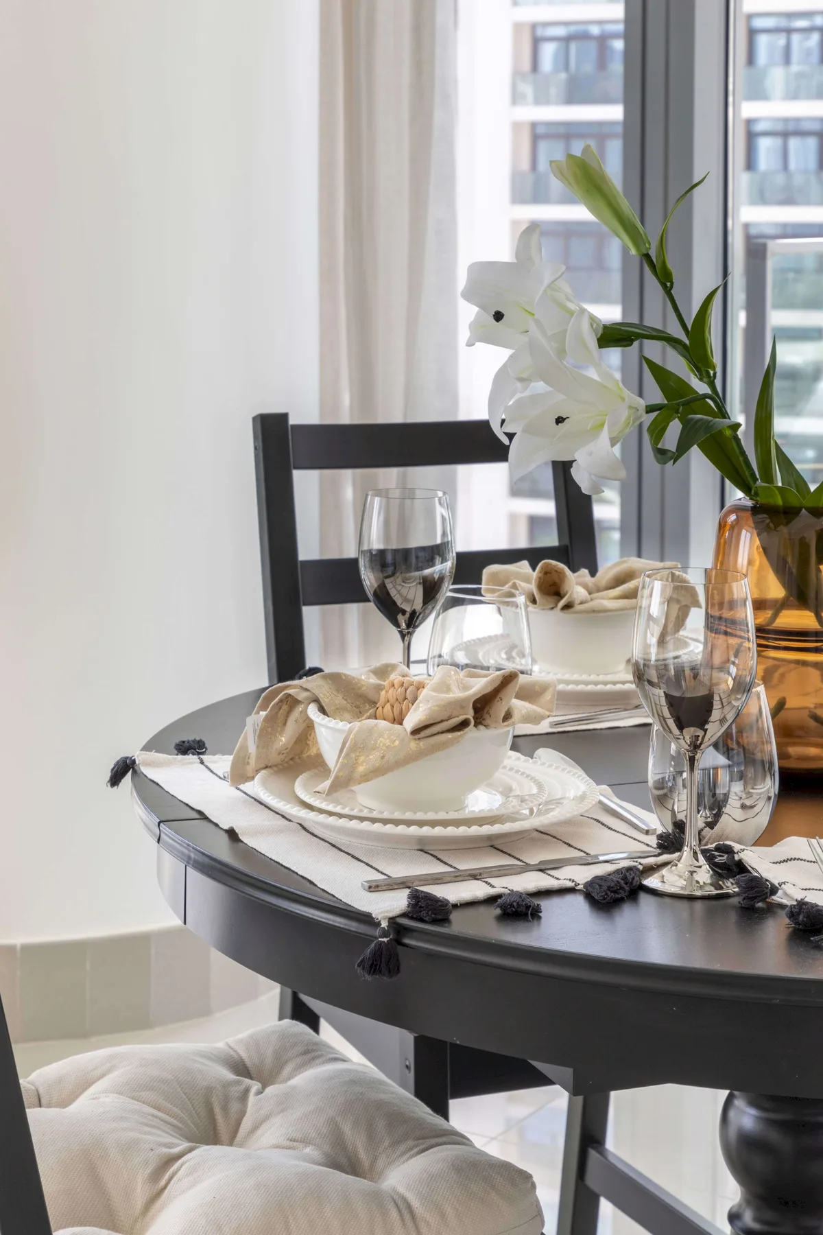 Una mesa de comedor está puesta con platos, copas y flores blancas junto a una ventana.