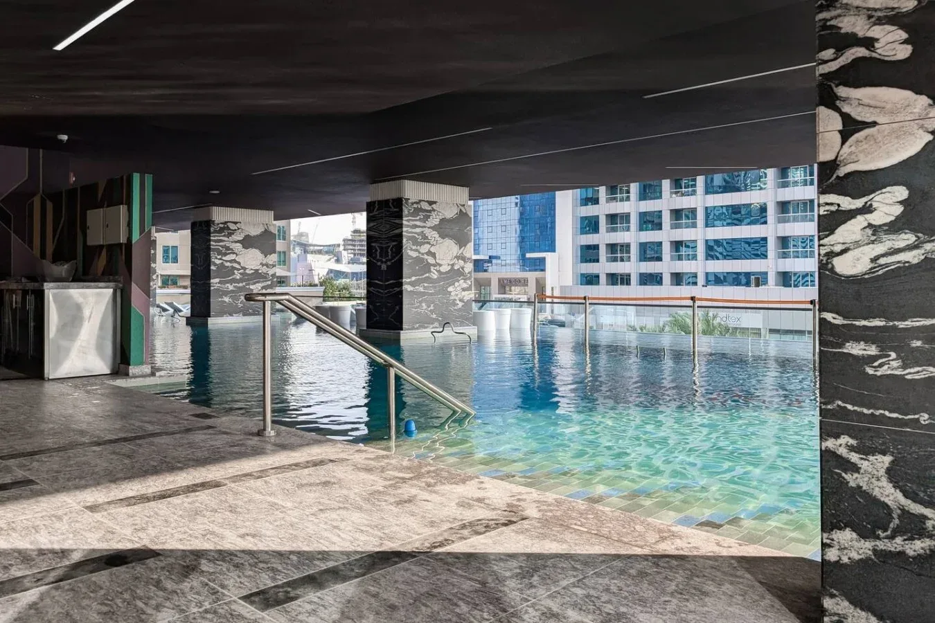 Una piscina está bajo una zona cubierta, con edificios urbanos visibles al fondo.