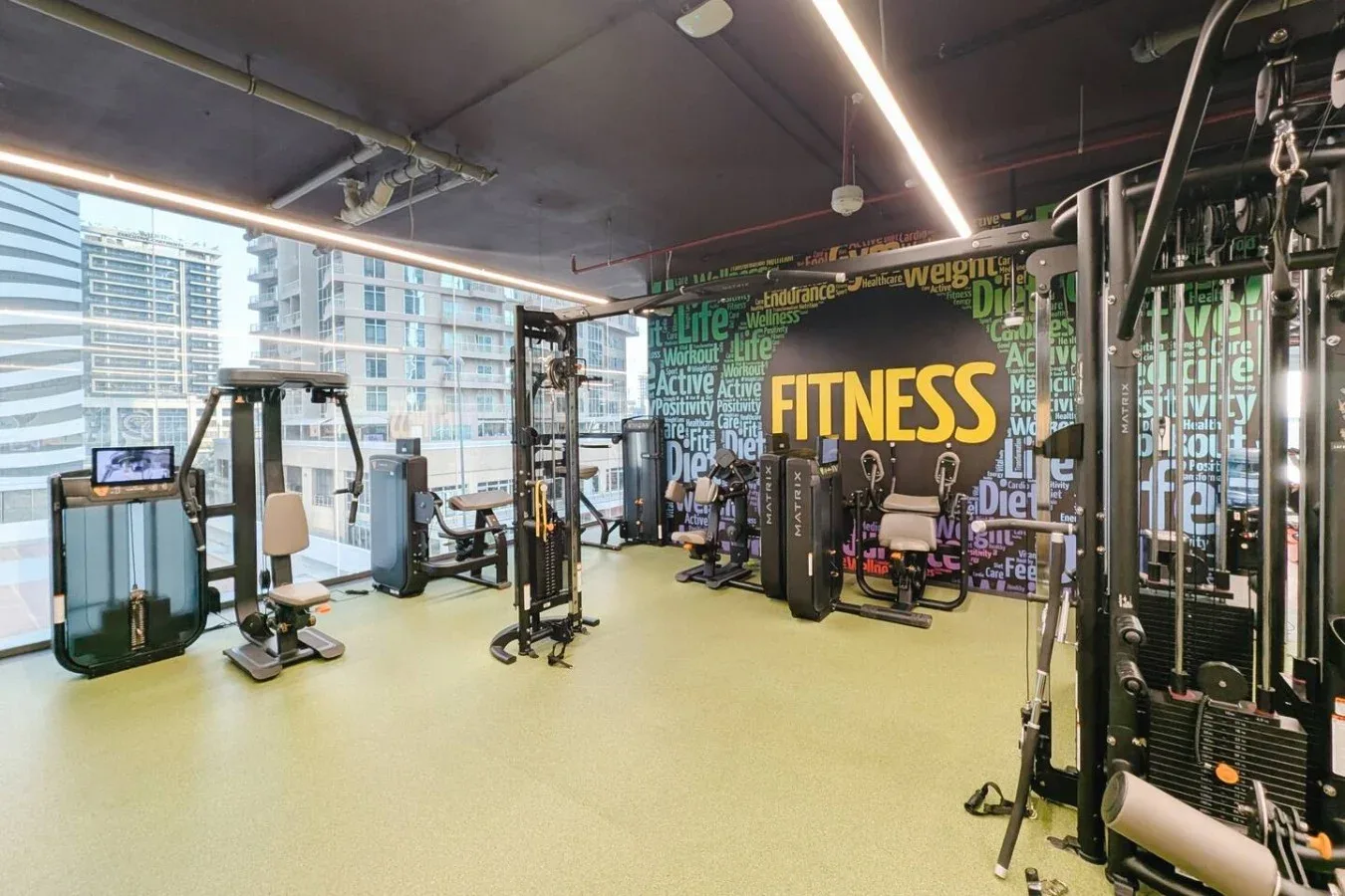Una sala de fitness con máquinas de ejercicio y grandes ventanas con edificios a la vista.