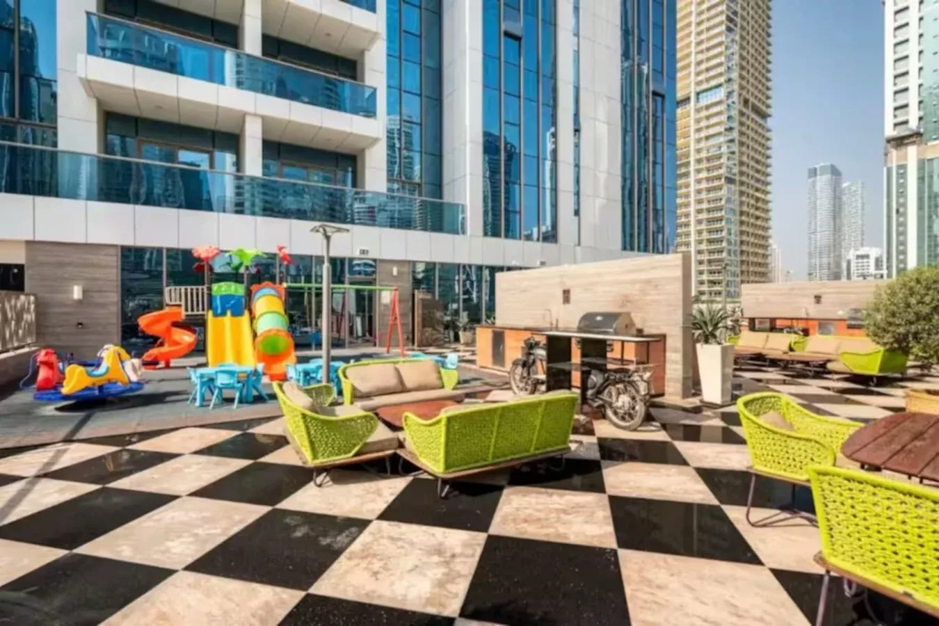 Una zona de descanso al aire libre tiene asientos verdes y una colorida estructura de juegos infantiles.