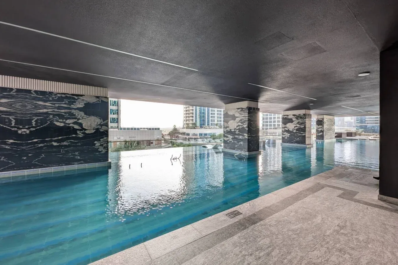 Una piscina cubierta bajo un techo de concreto, con edificios de la ciudad al fondo.