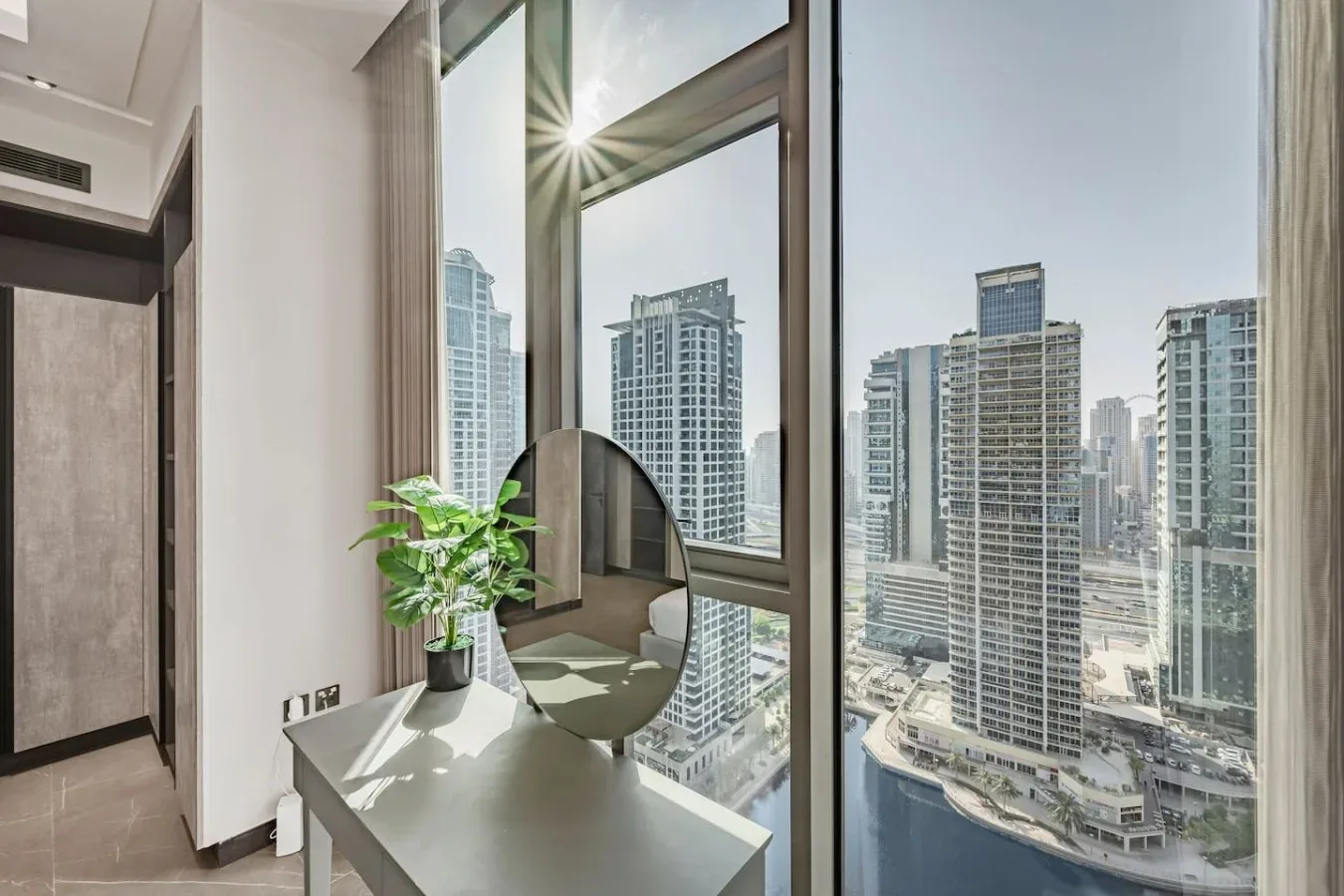 Una habitación luminosa tiene un escritorio y grandes ventanales con vistas a edificios de la ciudad.