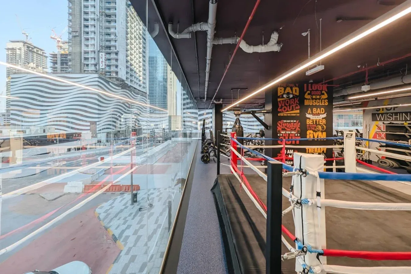 Un gimnasio de boxeo con un ring rodeado de cuerdas junto a grandes ventanas con vistas a edificios.