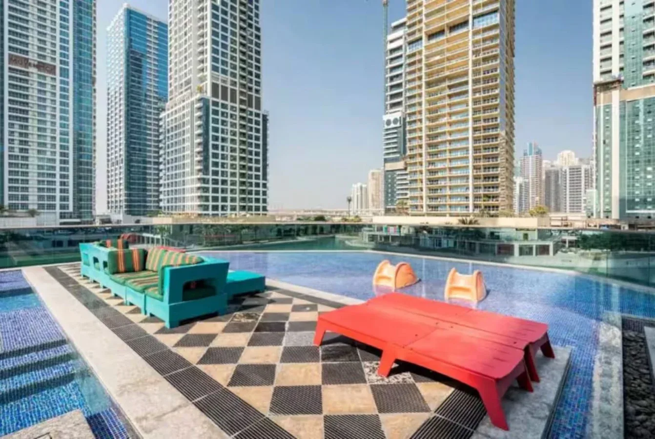 Una terraza con piscina, asientos turquesa y una mesa roja ofrece vistas de edificios altos.