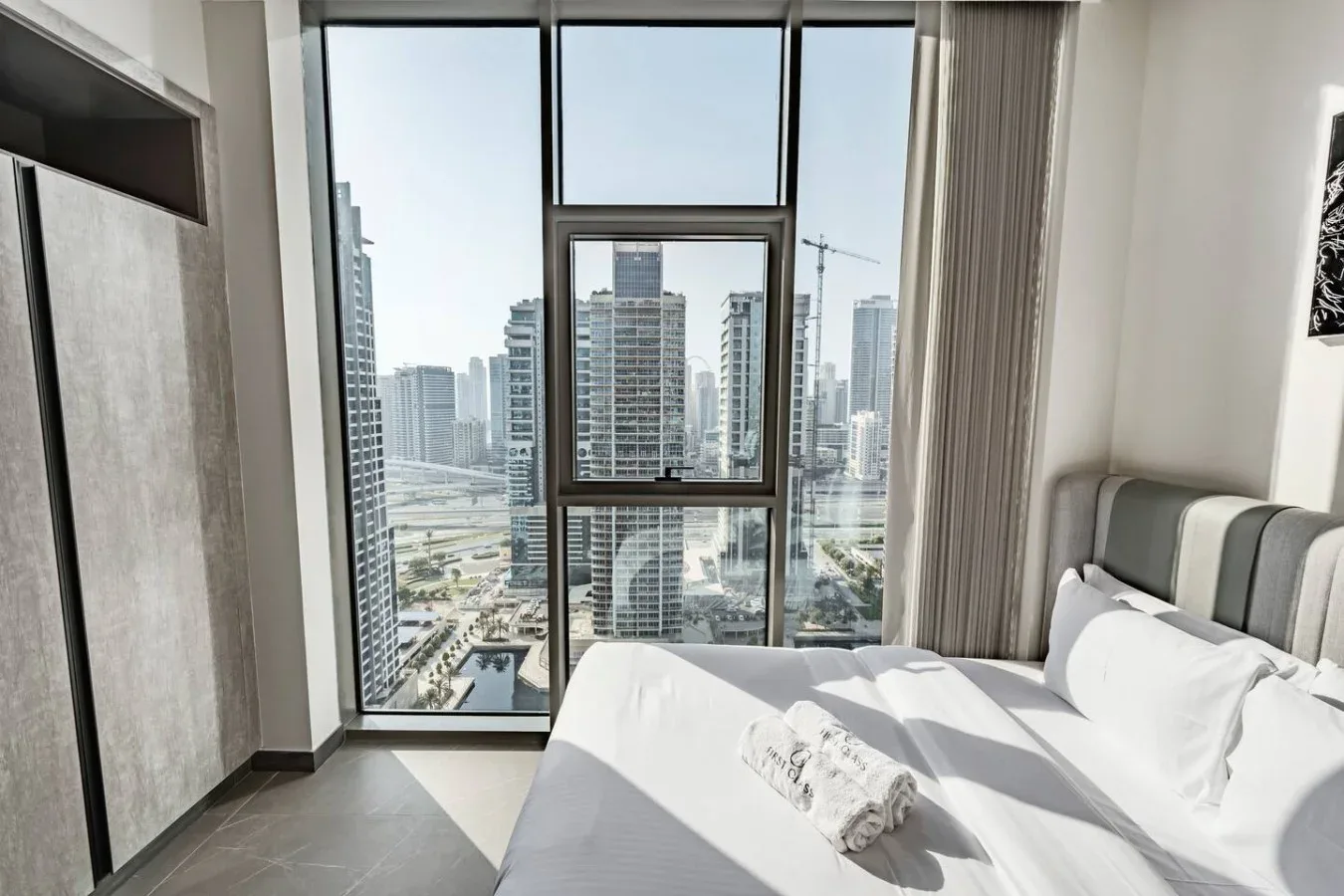 Un dormitorio luminoso con una cama junto a grandes ventanas con vistas a edificios de la ciudad.