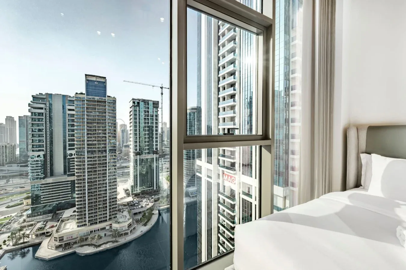 Un dormitorio con ropa de cama blanca junto a una ventana amplia con vistas a edificios.