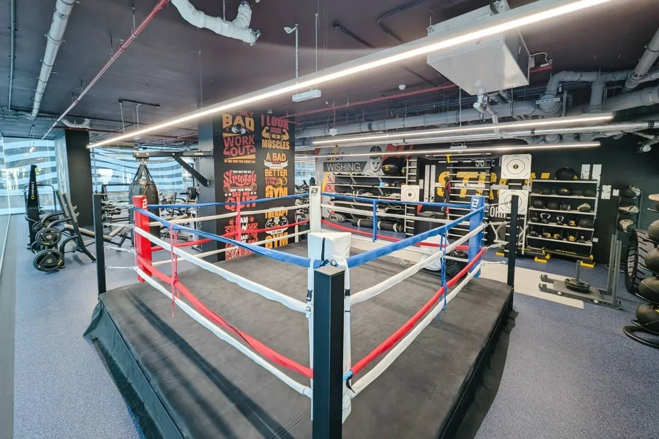 Un ring de boxeo está en un gimnasio rodeado de equipo de entrenamiento.