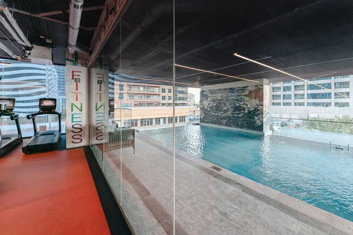 Una piscina cubierta junto a una zona de ejercicio, con grandes ventanales frente a edificios cercanos.