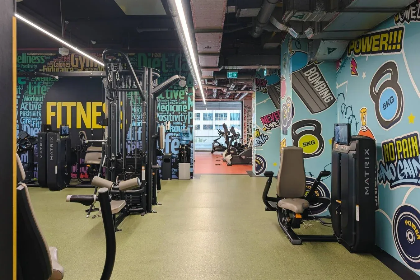 Interior de un gimnasio con máquinas de ejercicio y murales coloridos en las paredes.