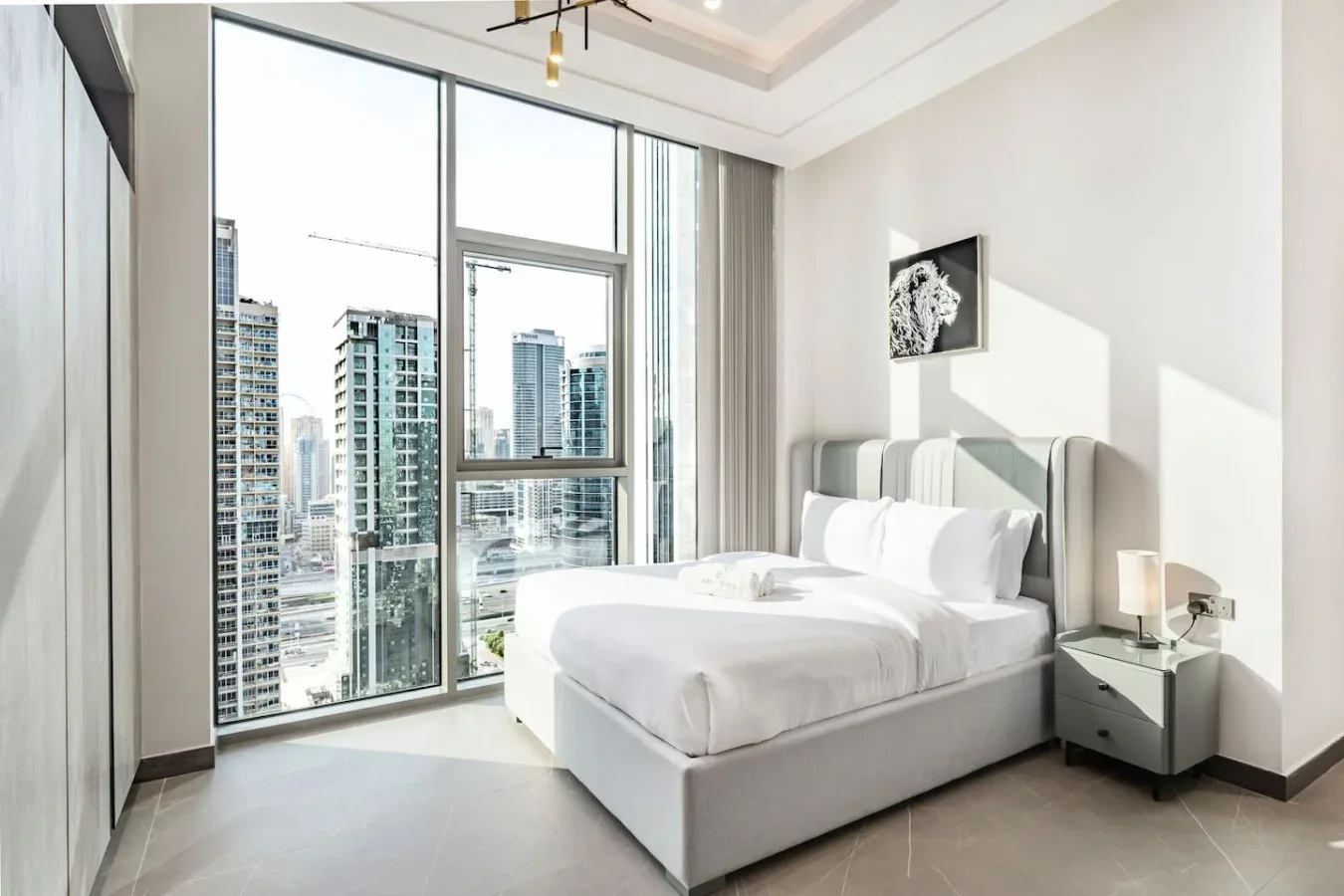 Un dormitorio luminoso con una cama junto a grandes ventanas con vistas a edificios de la ciudad.