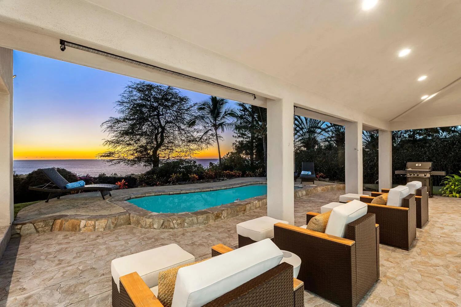 A covered outdoor seating area overlooks a pool and a sunset.