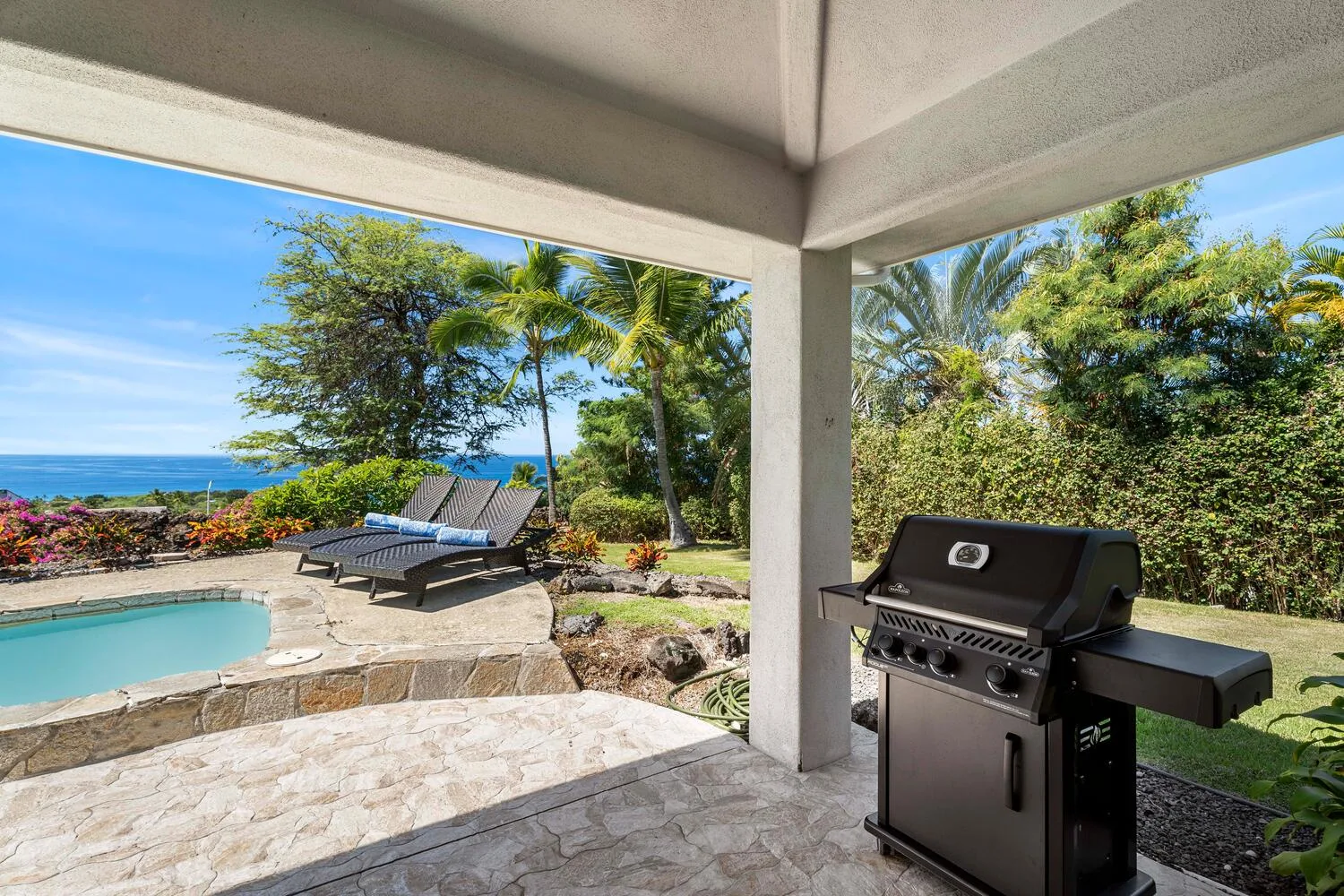 A covered patio overlooks a pool, lounge seating, garden greenery, and the ocean.