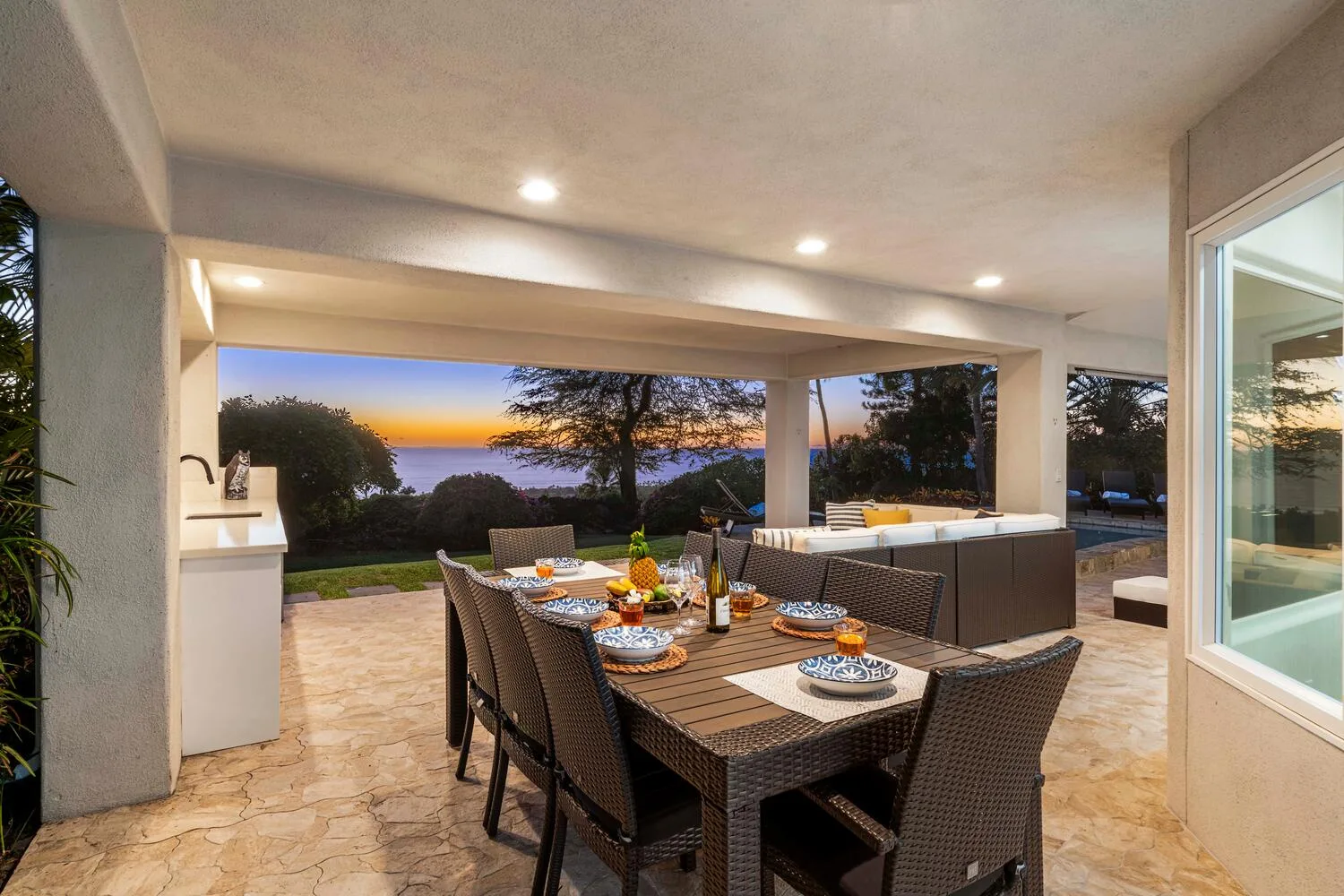 A covered outdoor dining area with a table and seating overlooking a garden at sunset.