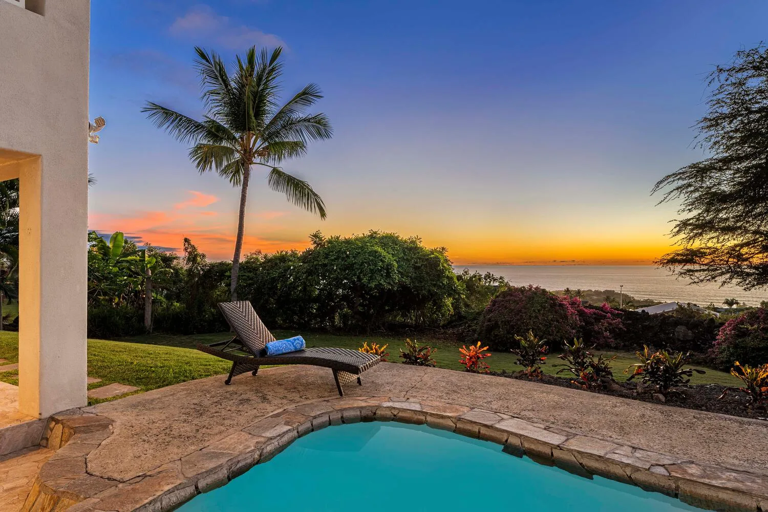 A swimming pool and patio overlook a landscaped garden and ocean at sunset.
