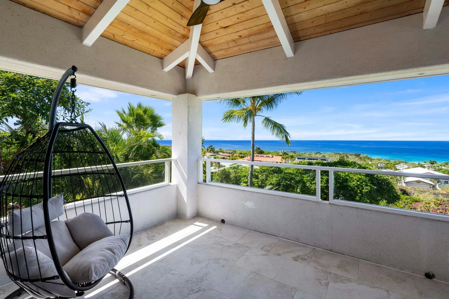 A covered balcony with a hanging chair overlooks tropical greenery and the ocean.