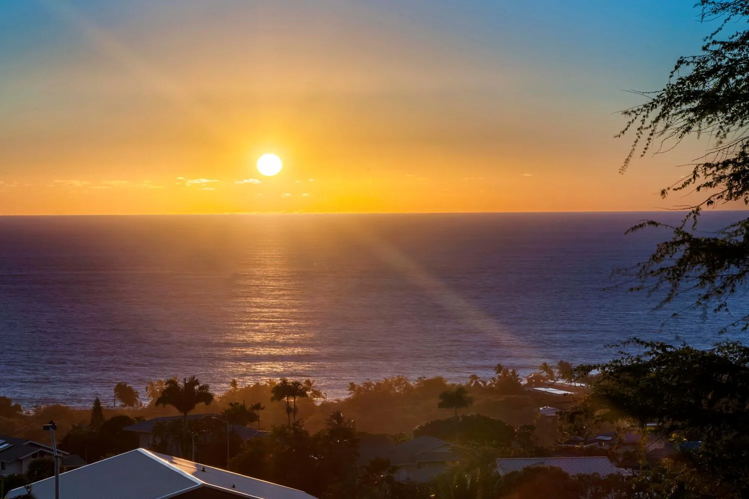 Sunset over the ocean, with rooftops and trees along the lower edge.