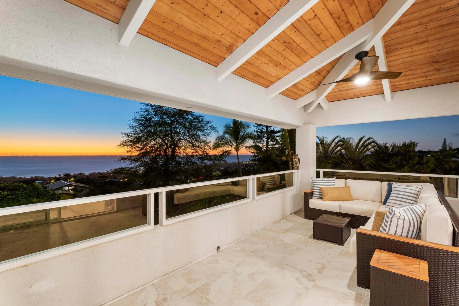 A covered terrace with cushioned seating overlooks trees and a sunset.