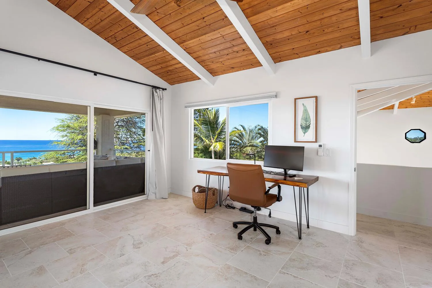 A bright room with a desk and seating beside wide windows overlooking the ocean.