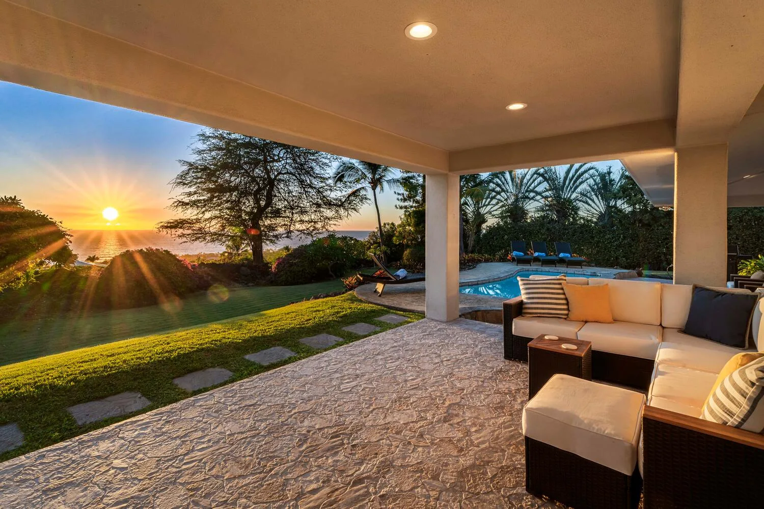 A covered patio with outdoor seating overlooks a pool and sunset.