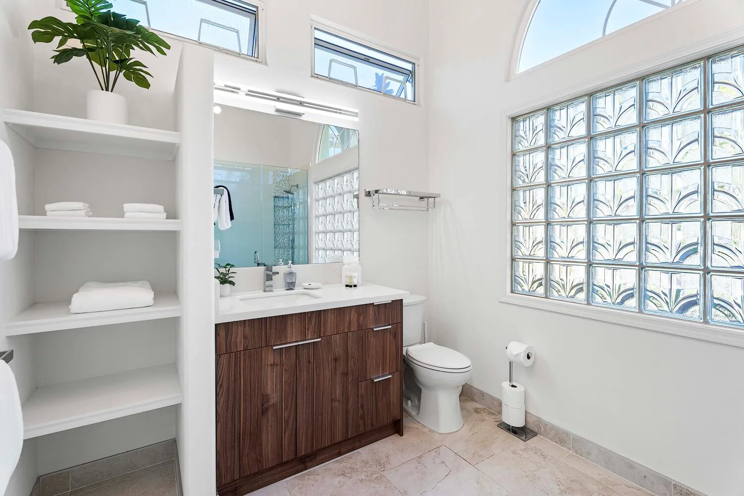 A bright bathroom with a wood vanity, toilet, and glass-block window.