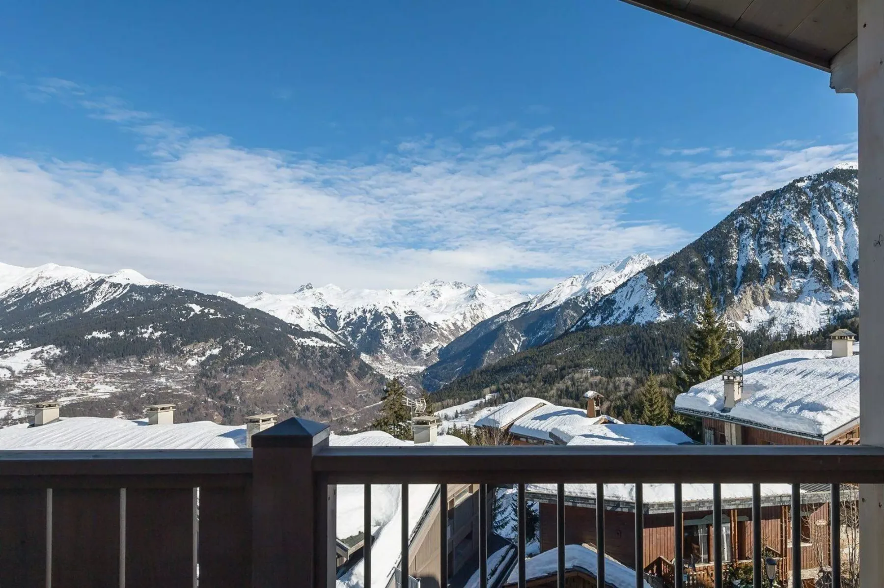 A balcony overlooks snow-covered rooftops and a mountain range beneath a blue sky.