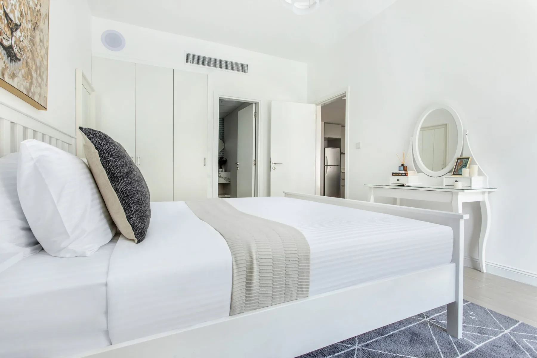 Un dormitorio luminoso con una cama blanca, ropa de cama gris y un tocador.