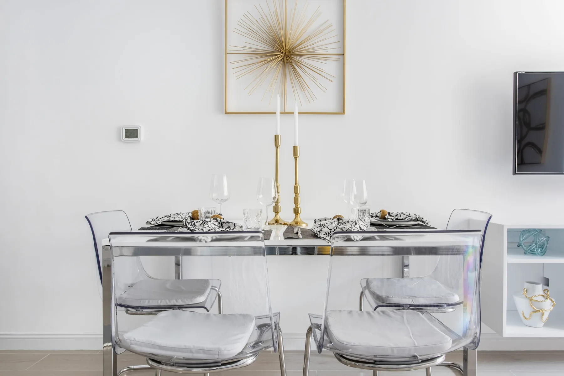Comedor con la mesa puesta y asientos transparentes bajo un cuadro enmarcado.
