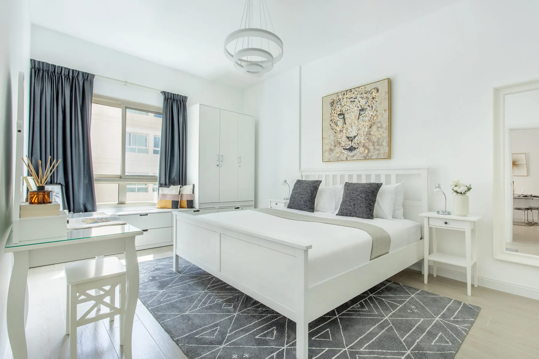 Un dormitorio luminoso con una cama blanca, una alfombra gris y un escritorio junto a la ventana.