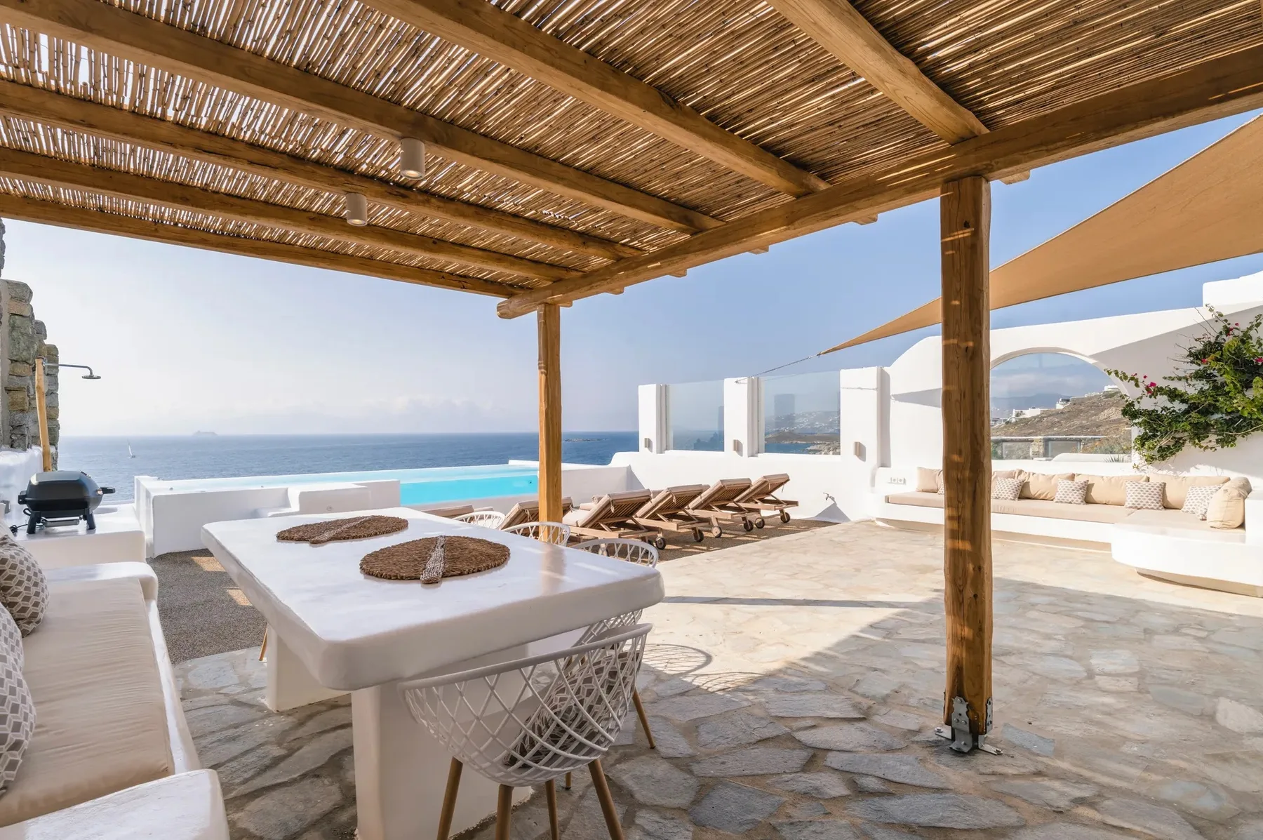 A seaside patio with a shaded dining table, lounge seating, and a view of the sea.