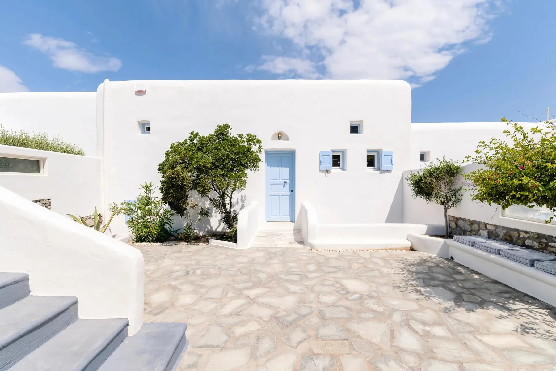 A white building with a blue door opens onto a sunny stone-paved courtyard.