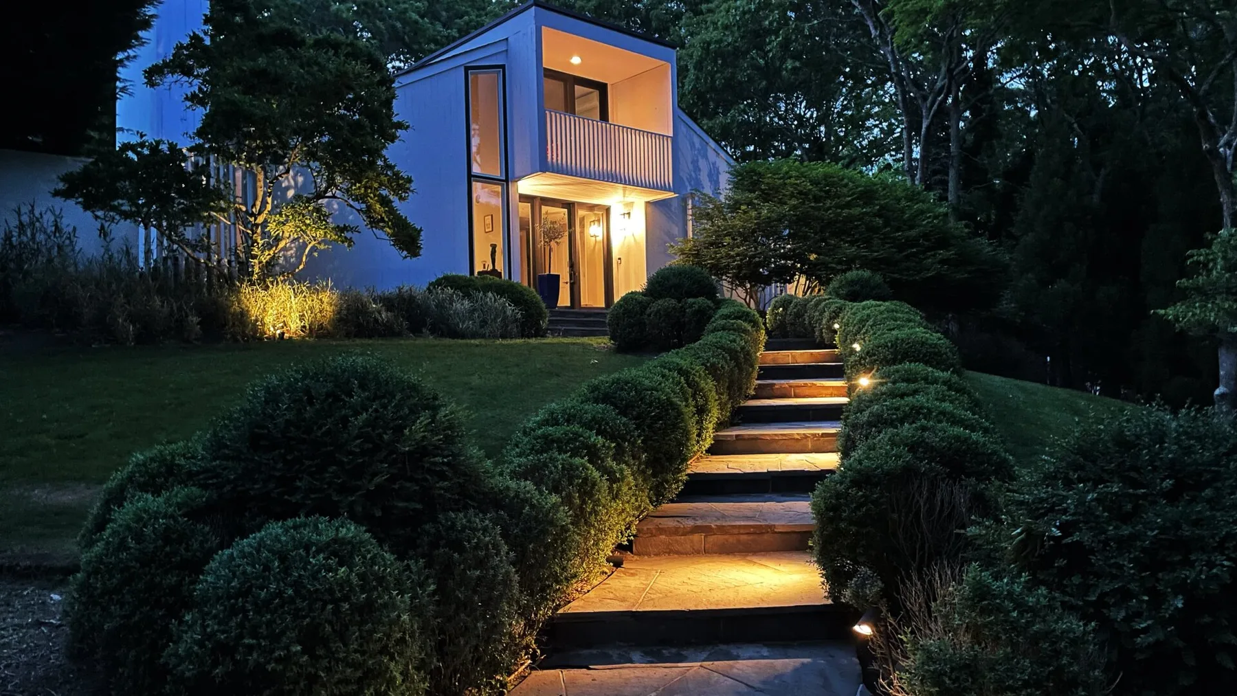 Una casa moderna brilla al anochecer junto a un camino ajardinado con escalones iluminados.