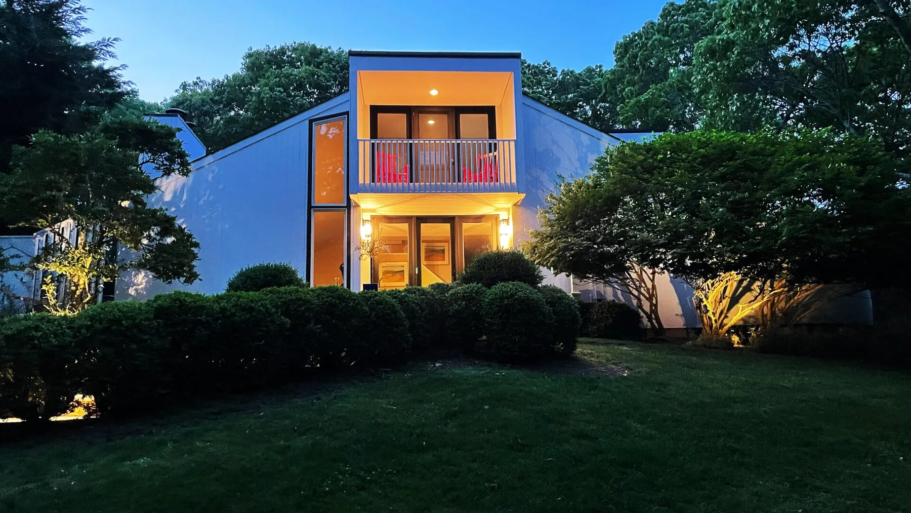 Una casa moderna se alza detrás de un seto, con luces cálidas al anochecer.