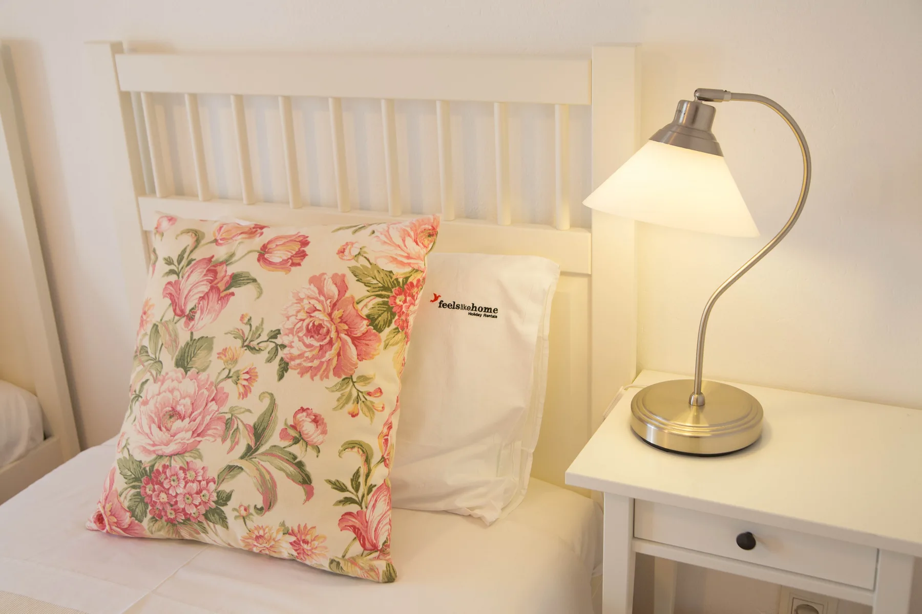 Un dormitorio con una cama blanca, un cojín floral y una lámpara de mesilla.
